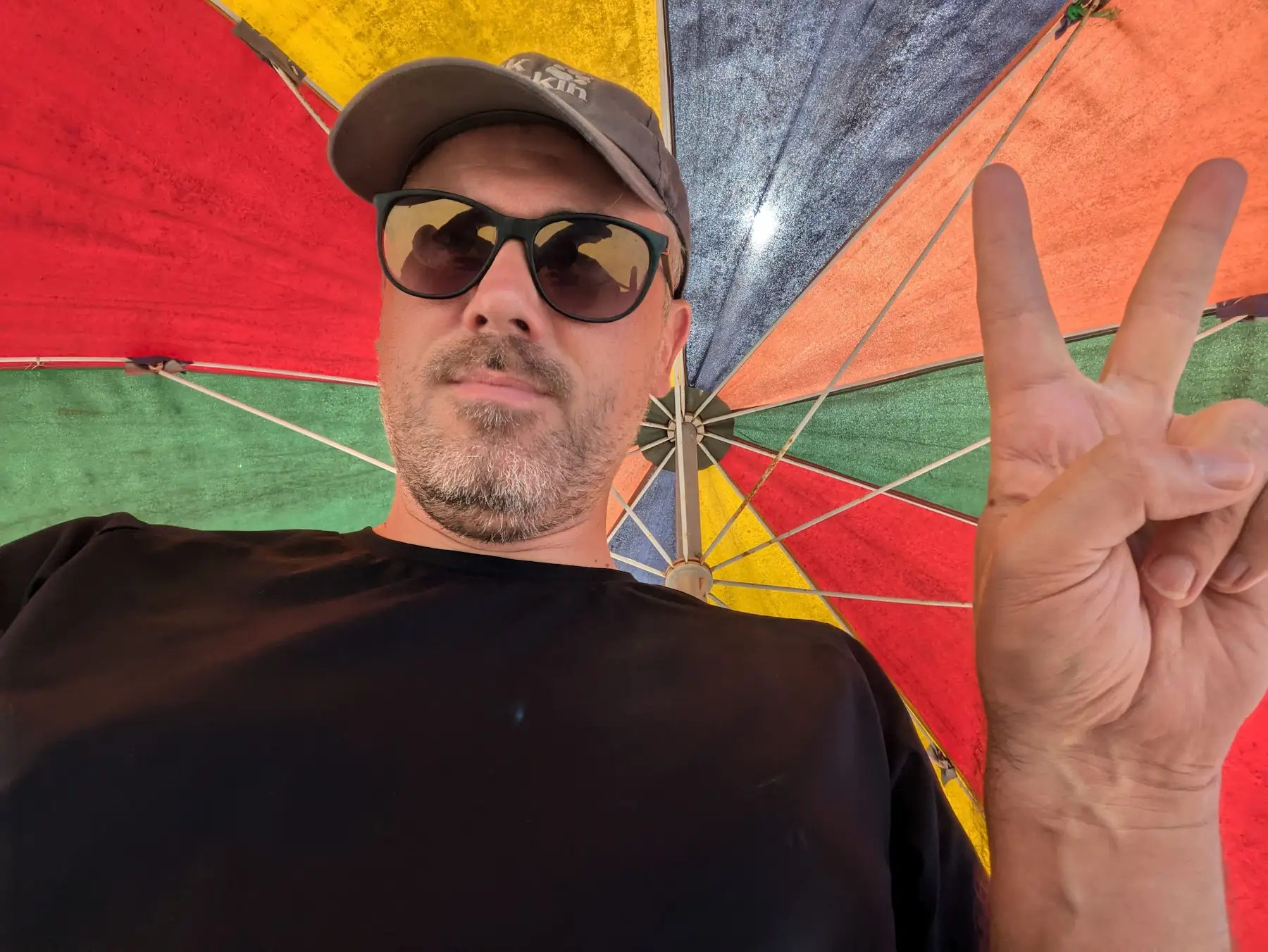 A man under a colourful umbrella making a peace sign.